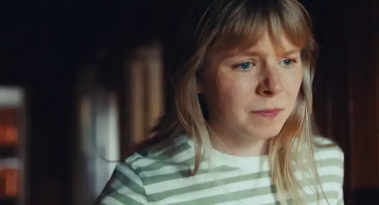 A woman with blonde hair and a striped shirt looks anxiously out of frame in a warmly lit room.