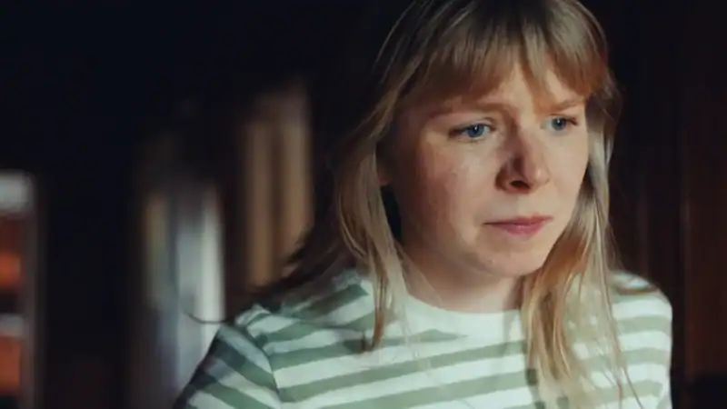 A woman with blonde hair and a striped shirt looks anxiously out of frame in a warmly lit room.
