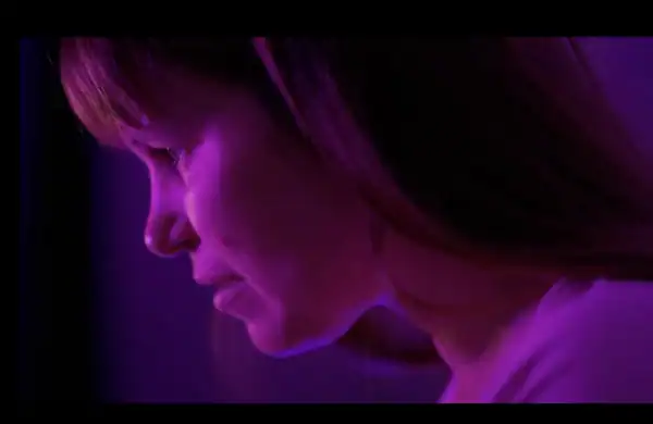 Close-up of a woman with shoulder-length hair illuminated by purple lighting, looking downward with a contemplative expression.