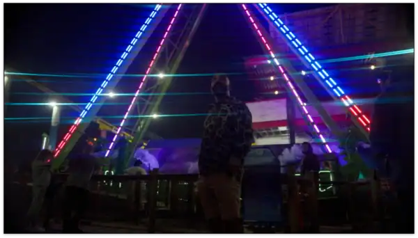 Person standing in front of colorful neon light installation at a nighttime event or amusement park.