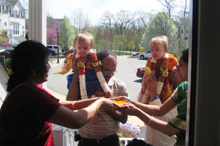 A group of adults stand at the entrance of a house, holding two toddlers dressed in festive clothing and garlands. The adults appear to be performing a traditional Indian welcoming ceremony, offering a plate with colored powders and fruit (bananas) as part of the ritual. Blossoming trees and residential houses are visible in the background on a sunny day.