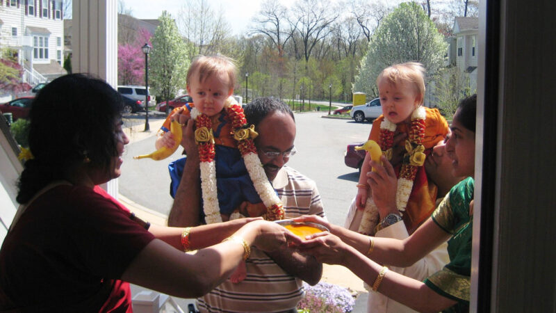 A group of adults stand at the entrance of a house, holding two toddlers dressed in festive clothing and garlands. The adults appear to be performing a traditional Indian welcoming ceremony, offering a plate with colored powders and fruit (bananas) as part of the ritual. Blossoming trees and residential houses are visible in the background on a sunny day.