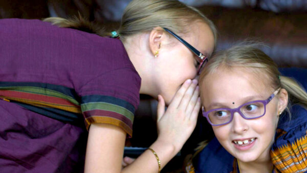 A girl wearing glasses whispers into the ear of another girl with glasses, who is smiling and listening.