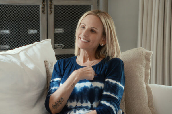 A woman with shoulder-length blonde hair sits on a couch, smiling and signing. She wears a blue and white tie-dye top and has a visible tattoo on her forearm.