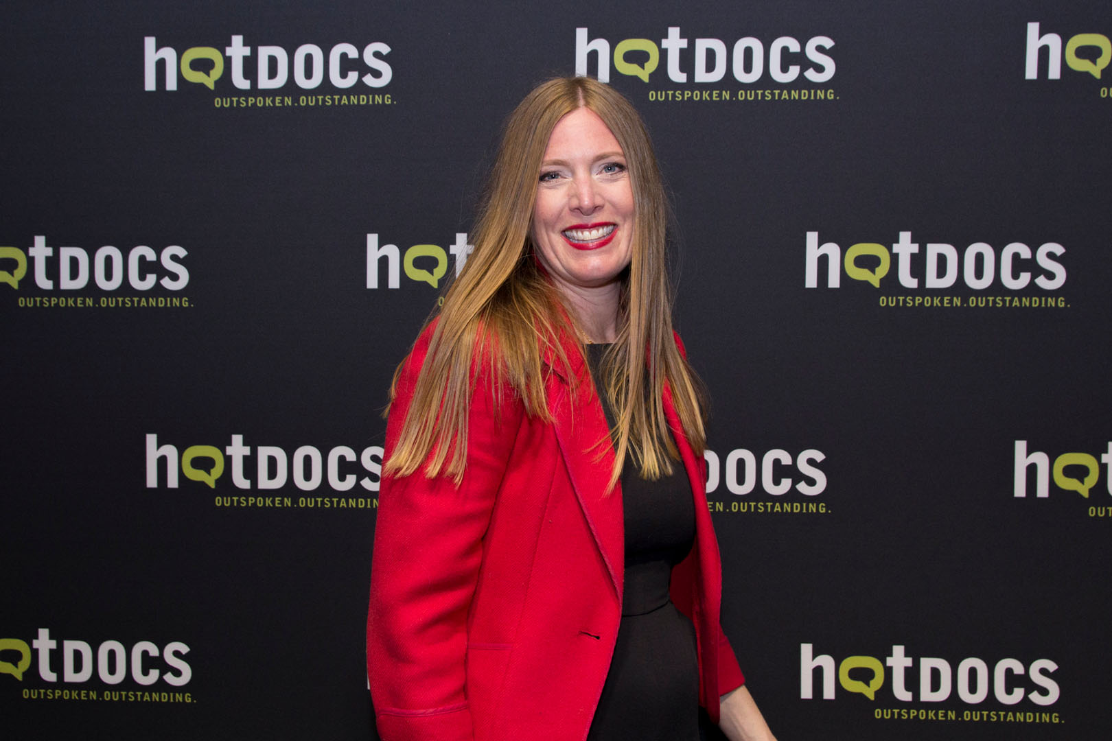 A person with long blonde hair stands in front of a repeating "hotdocs" festival backdrop, wearing a black top and a bold red jacket. The setting is a media wall at a film event, conveying a professional yet lively atmosphere associated with documentary screenings and industry gatherings.