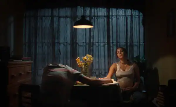 A woman sitting at a dining table with a man, holding her pregnant belly, in a dimly lit room with blue curtains and a hanging lamp, conveying a moment of reflection or anticipation.