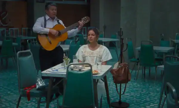 A man playing guitar and a woman sitting at a table in a restaurant or cafe setting with empty chairs and tables around them.
