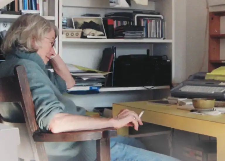 Woman with gray hair and glasses sitting at a cluttered desk in a home office with shelves of books and CDs in the background.