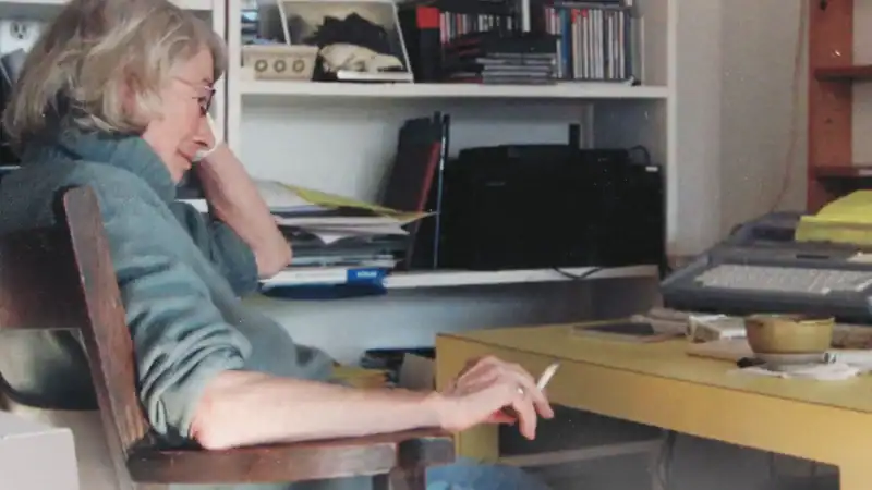 Woman with gray hair and glasses sitting at a cluttered desk in a home office with shelves of books and CDs in the background.