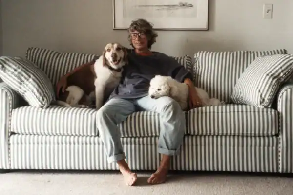 Person sitting on a striped sofa with two dogs, one sitting beside and one lying on their lap, in a cozy living room setting.