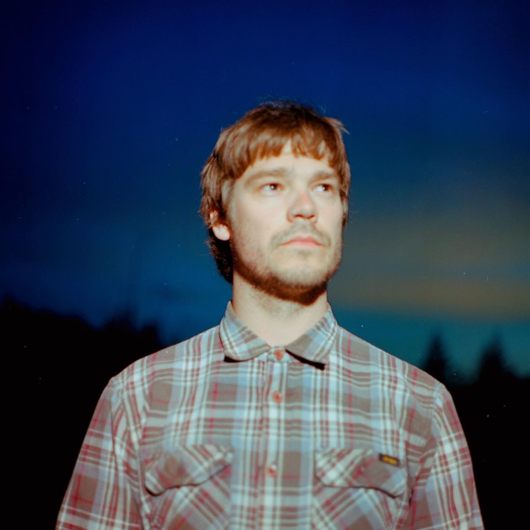 A person stands outdoors at dusk, wearing a plaid button-up shirt. The sky behind them is deep blue with a faint sunset glow near the horizon, and silhouettes of trees are visible in the distance. The lighting and colors evoke a tranquil, contemplative atmosphere.
