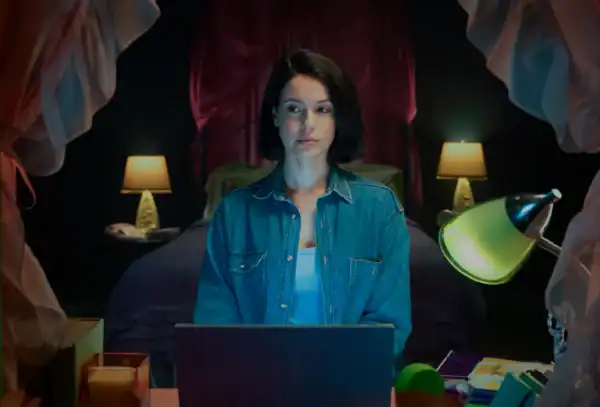 Woman sitting with a laptop at a desk with colorful objects, in a dimly lit room with bedside lamps.