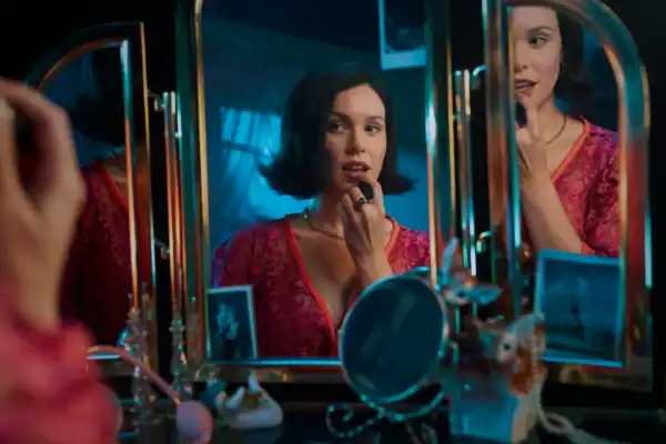 A woman with short dark hair applying lipstick while looking into a mirror surrounded by makeup and beauty products in a dimly lit room.