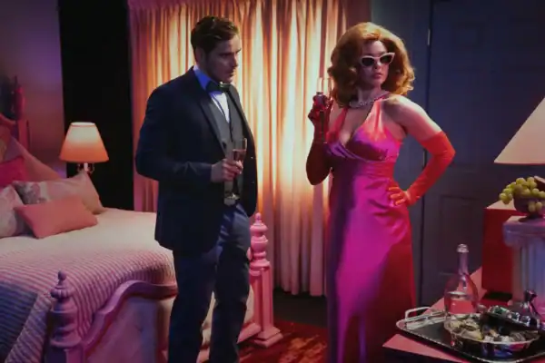 A man in a tuxedo and a woman in a pink satin dress with sunglasses stand in a dimly lit bedroom with vintage decor, holding drinks and engaging in conversation.