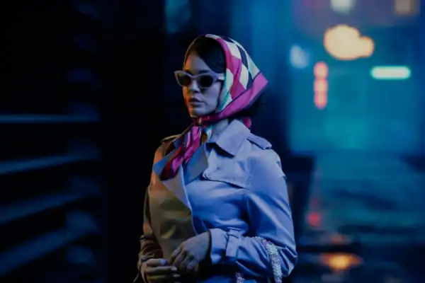 Stylish woman wearing sunglasses and a colorful headscarf walking in an urban night scene with neon lights.