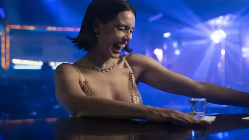 A woman with short dark hair smiling and reaching out with her arm in a vibrant, colorful nightclub or concert setting with blue and purple lighting.