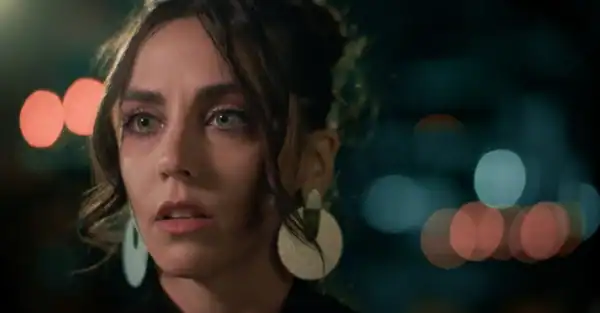 Close-up of a woman with dark hair and earrings, illuminated by city lights at night, emphasizing her facial features and contemplative expression.