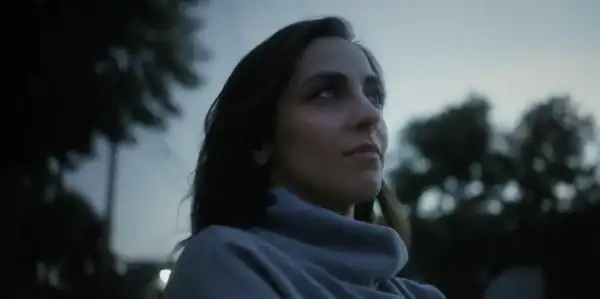 A woman with dark hair and a contemplative expression outdoors during dusk, with trees and a cloudy sky in the background.