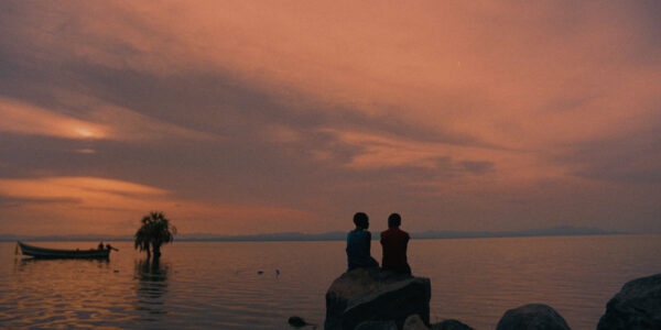 Two people sitting on a rock in he distance, facing the sea at sunset.