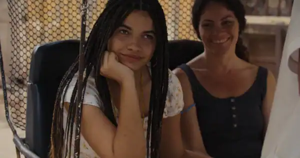 Young woman with braided hair sitting at a table with a smiling woman in a black top, in a casual indoor setting with a wire fence background.