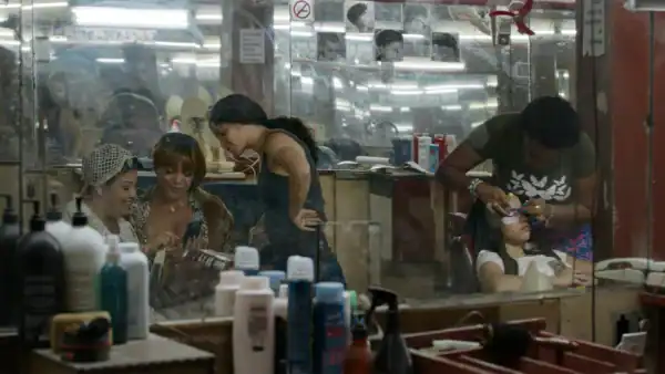 People getting haircuts and styling at a busy barbershop with mirrors, hair products, and salon tools visible in the background.