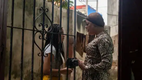 Two women engaged in a conversation behind a metal gate, one with long braided hair and the other wearing a hat, in an outdoor setting with a weathered wall and hanging cloths.