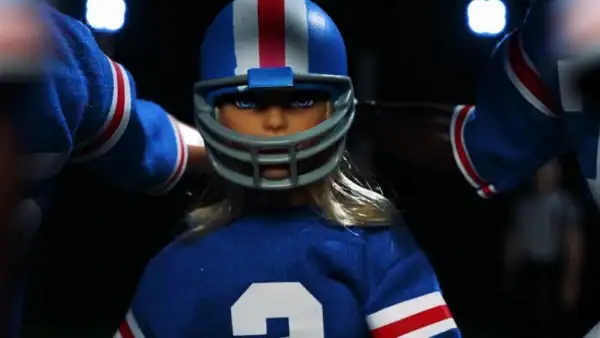 A blonde doll wearing a football helmet and jersey with the number 2, ready for a game, in a sports setting with bright lights in the background.
