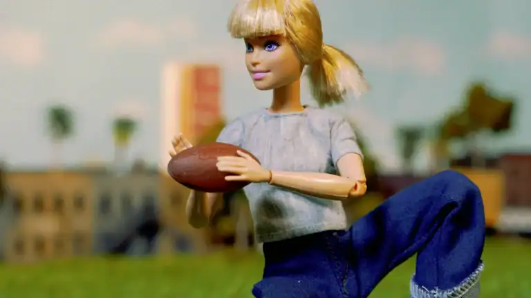 A doll with blonde hair, wearing a gray shirt and blue jeans, holding a football in an outdoor setting with a cityscape background.