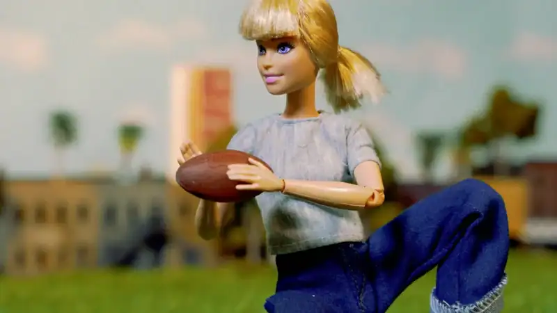 A doll with blonde hair, wearing a gray shirt and blue jeans, holding a football in an outdoor setting with a cityscape background.