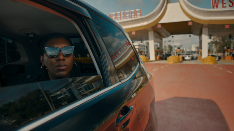 Person with sunglasses looks out from a car window, reflecting urban landscape.