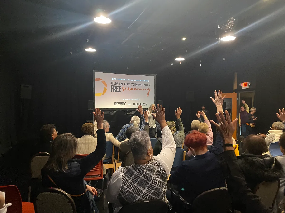 A group of people seated in a dark room raise their hands while facing a screen displaying "Film in the Community Free Screening.