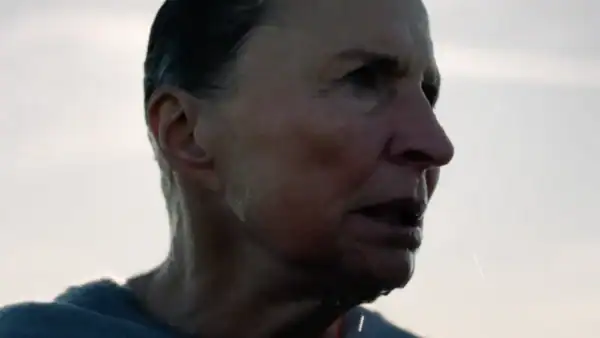 Close-up of a woman with short dark hair and a serious expression, outdoors with a cloudy sky background.