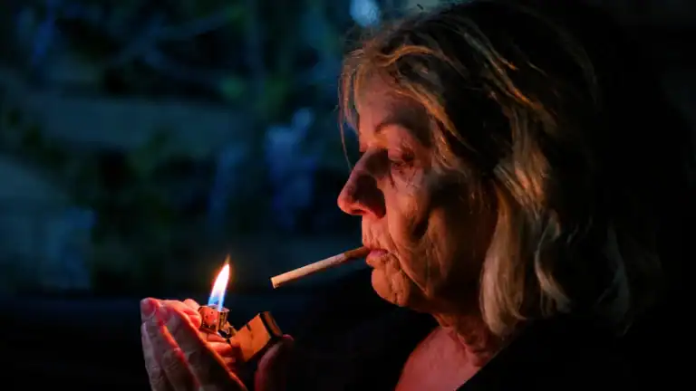 Close-up of an elderly woman lighting a cigarette with a lighter in a dark setting, highlighting her profile and detailed facial features.