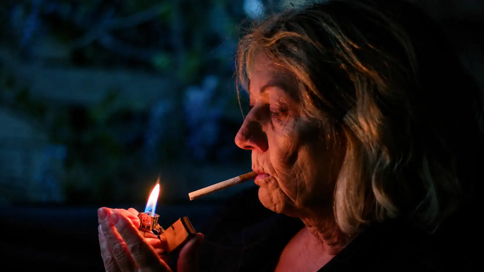 Close-up of an elderly woman lighting a cigarette with a lighter in a dark setting, highlighting her profile and detailed facial features.