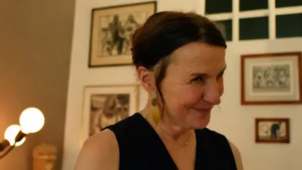 Woman with dark hair and earrings smiling in a warmly lit room with framed pictures on the wall.
