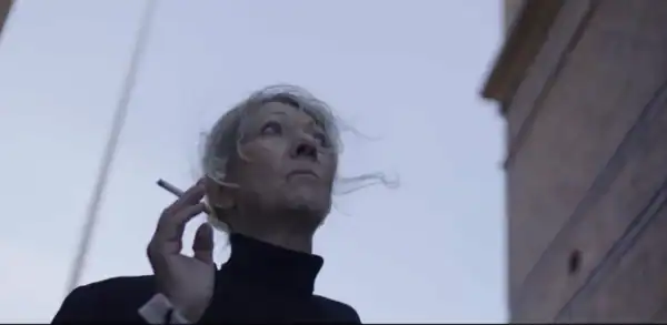 A woman with gray hair and a black turtleneck holds a cigarette near her face, looking upward against a cloudy sky and historic building background.