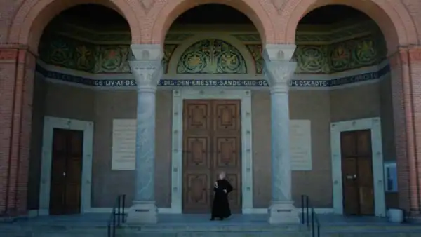 A person in black clothing stands in front of a historic building with ornate columns and decorative friezes, featuring a large wooden door and inscriptions on the walls.