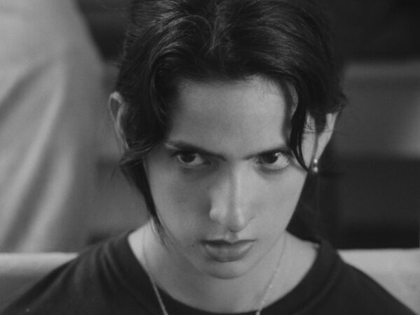 Black and white close-up of a young person with dark hair, looking intensely forward with a serious, almost stern expression. They wear a dark shirt and necklace, with loose strands of hair framing their face.