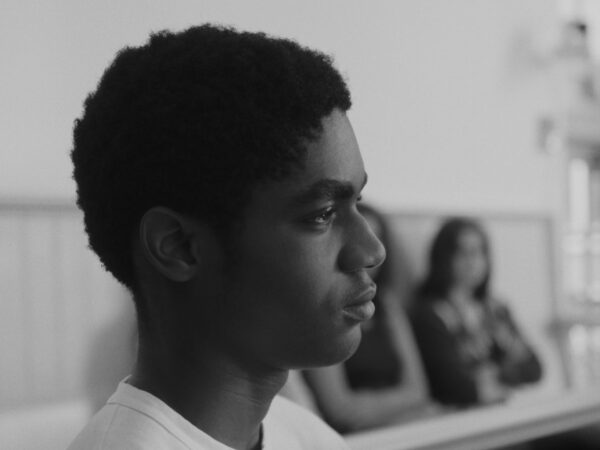 Black and white photo of a young man in profile, facing right, with a serious expression. He wears a light-colored shirt and sits indoors against a paneled wall. In the blurred background, two women are seated, out of focus.