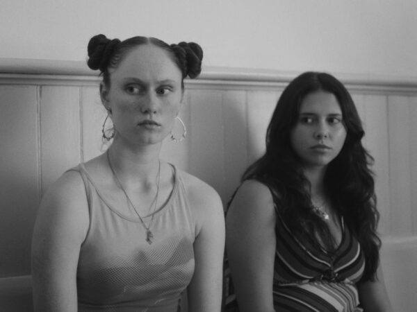 Black and white photo of two young women sitting side by side on a bench against a paneled wall. The woman on the left has her hair in two braided buns, wearing large hoop earrings and a sleeveless top, looking slightly to her left. The woman on the right has long dark hair, wearing a striped sleeveless top, and is also looking left, with a serious expression.