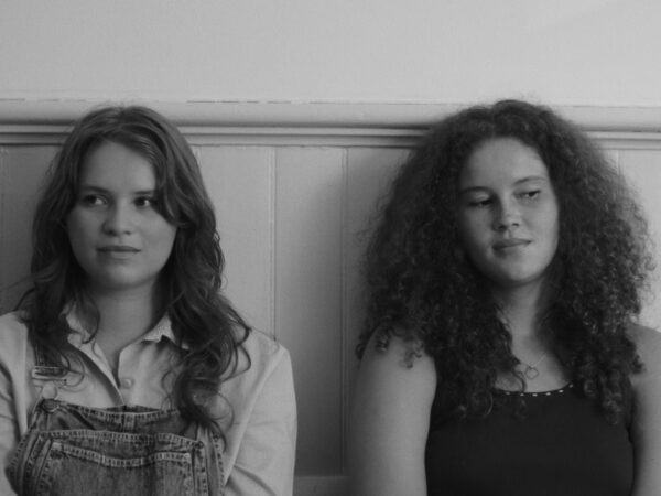 Black-and-white close-up of two teenage girls sitting side by side against a wooden paneled wall. The girl on the left wears denim overalls and a button-up shirt, looking slightly to the side. The girl on the right has curly hair, wears a tank top, and glances at the other girl with a subtle expression. The mood appears contemplative or quietly expressive.