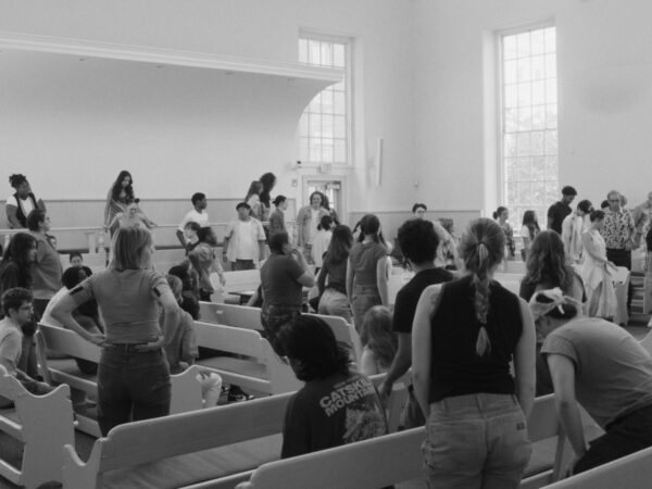A black-and-white photo of a large group of people, mostly teenagers, gathered in a bright, spacious room with high ceilings and large windows. They are standing or sitting on white wooden pews arranged in rows, suggesting a meeting or assembly in a place resembling a chapel or community hall. The atmosphere appears informal and social.