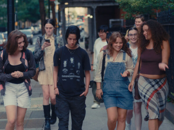 A diverse group of teenagers walk together on a city sidewalk, chatting and smiling. The group includes both boys and girls dressed in casual, expressive clothing like overalls, graphic tees, plaid skirts, and backpacks. One person is looking at a phone, while others are engaged in conversation. The background shows a tree-lined urban street with buildings and parked cars.