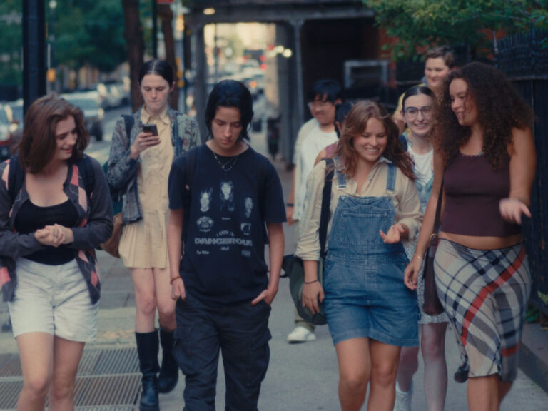 A diverse group of teenagers walk together on a city sidewalk, chatting and smiling. The group includes both boys and girls dressed in casual, expressive clothing like overalls, graphic tees, plaid skirts, and backpacks. One person is looking at a phone, while others are engaged in conversation. The background shows a tree-lined urban street with buildings and parked cars.