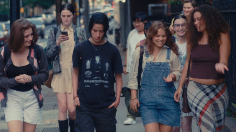 A diverse group of teenagers walk together on a city sidewalk, chatting and smiling. The group includes both boys and girls dressed in casual, expressive clothing like overalls, graphic tees, plaid skirts, and backpacks. One person is looking at a phone, while others are engaged in conversation. The background shows a tree-lined urban street with buildings and parked cars.
