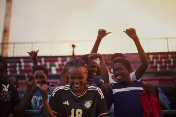Women soccer players throw up hands in celebration with stadium bleachers behind them