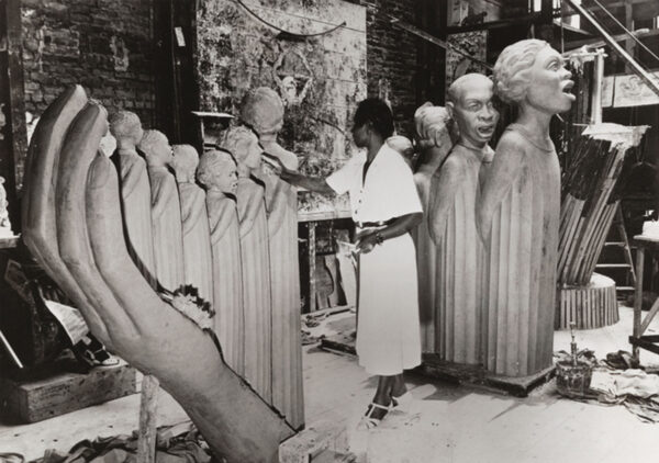 Black and white photograph of artist Augusta Savage in her studio, sculpting her monumental work The Harp. She stands in a white dress, shaping one of the many figures that form the strings of the harp-like sculpture. Surrounding her are large sculpted heads and figures, as well as studio tools and materials.