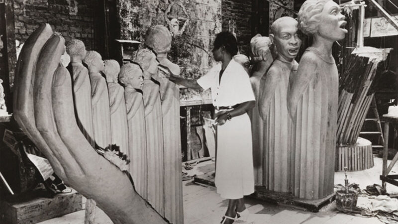 Black and white photograph of artist Augusta Savage in her studio, sculpting her monumental work The Harp. She stands in a white dress, shaping one of the many figures that form the strings of the harp-like sculpture. Surrounding her are large sculpted heads and figures, as well as studio tools and materials.