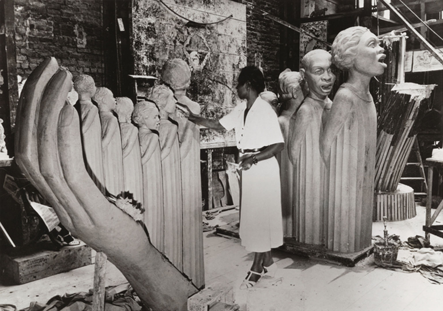 Black and white photograph of artist Augusta Savage in her studio, sculpting her monumental work The Harp. She stands in a white dress, shaping one of the many figures that form the strings of the harp-like sculpture. Surrounding her are large sculpted heads and figures, as well as studio tools and materials.