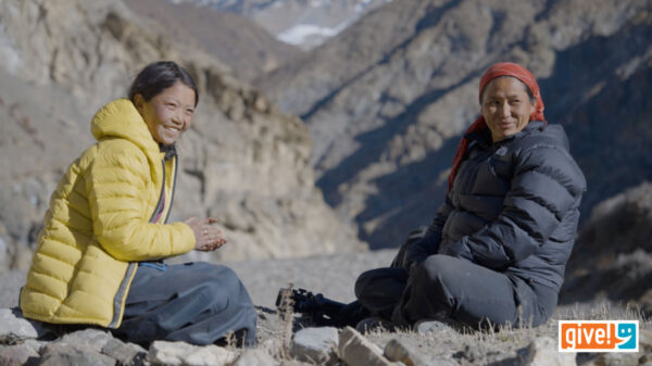 Two women wearing outdoor jackets sit on rocky ground in a mountainous landscape, smiling and facing the camera. The "give!" logo appears in the bottom right corner.